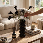 Hands arranging calla stems in a black bobbin vase on a rustic table before a cream sofa