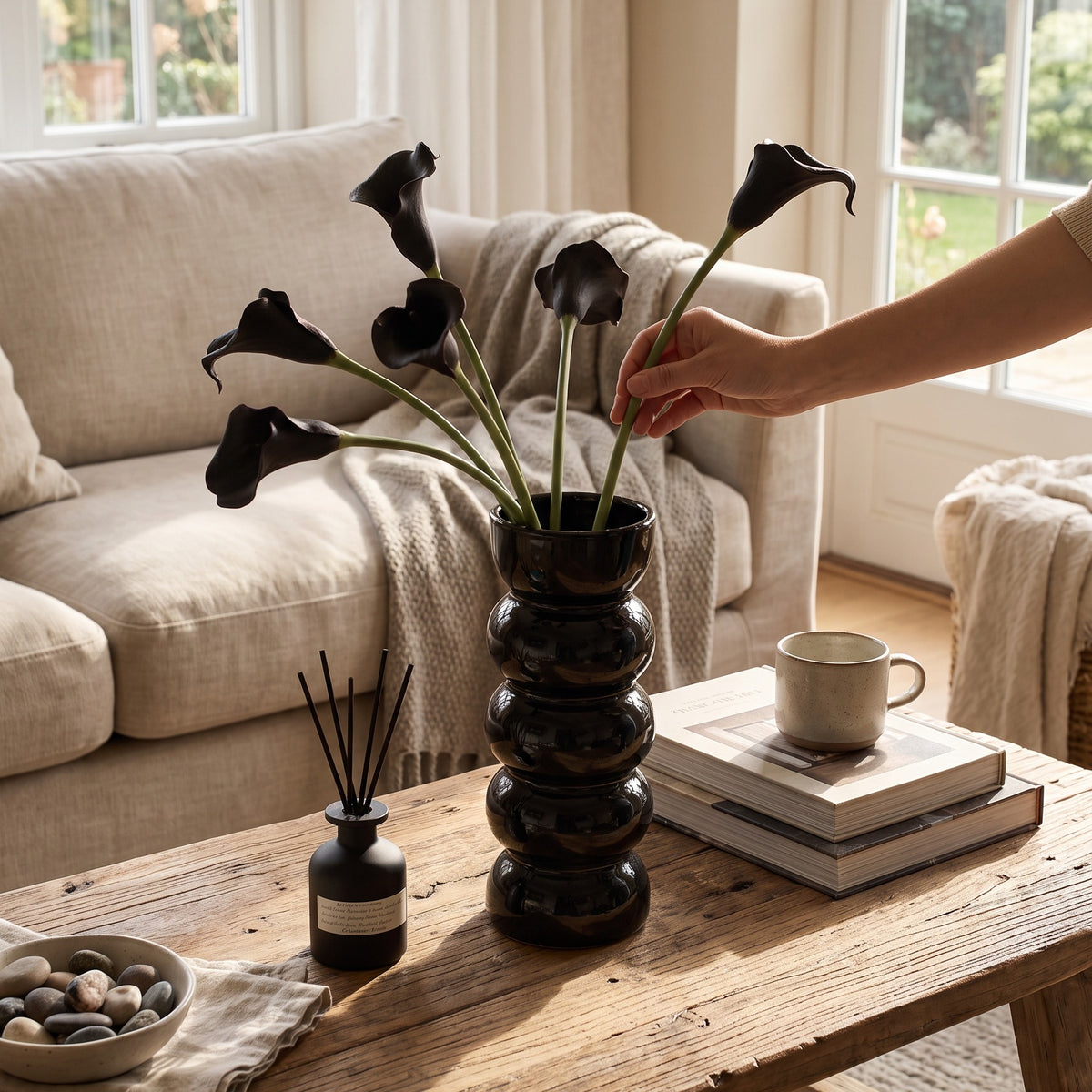 Hands arranging calla stems in a black bobbin vase on a rustic table before a cream sofa