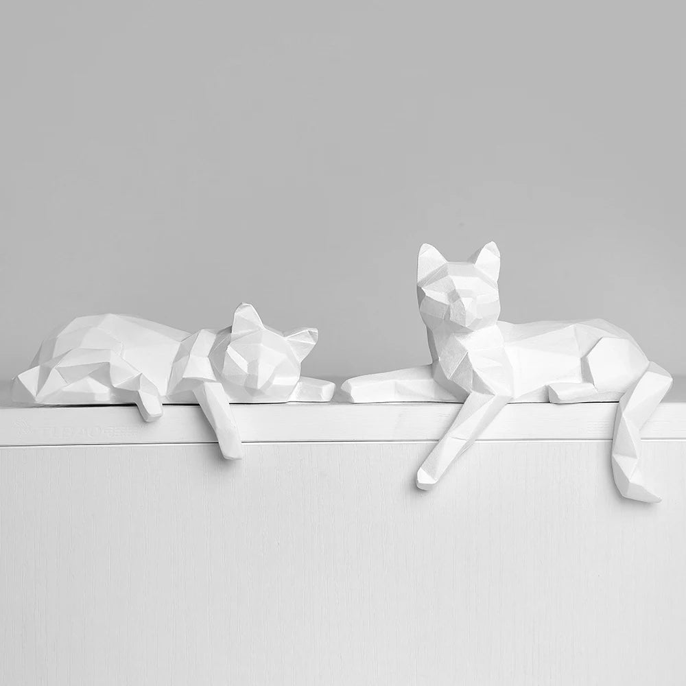 Two white faceted cats stretched out on a white ledge against a soft gray wall