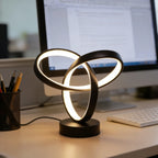 Black triple loop knot lamp lit on a desk beside a monitor and a pen cup