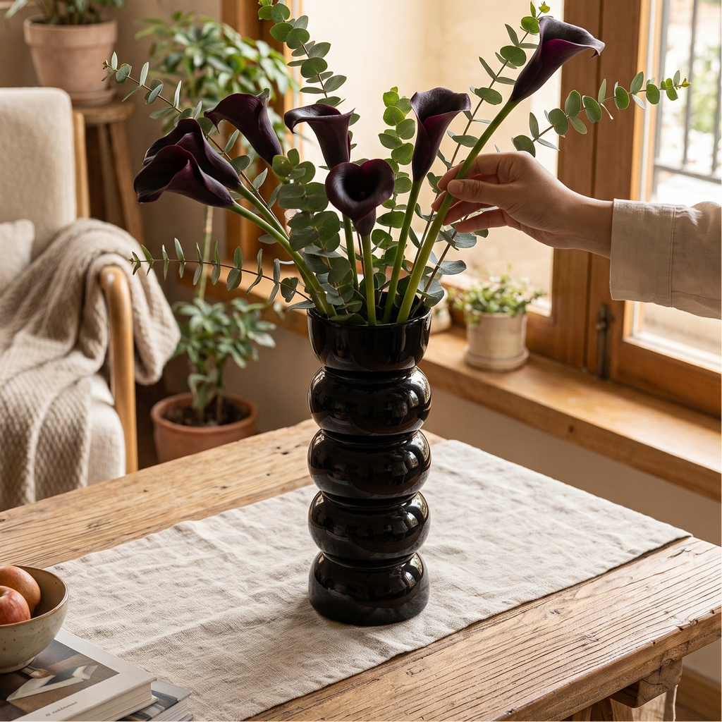 Glossy black stacked ball vase of dark calla lilies on a wooden table by a window