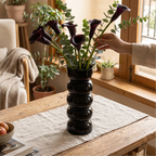 Glossy black stacked ball vase of dark calla lilies on a wooden table by a window