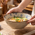 Hands carrying a floral bowl of noodle soup over a wooden table