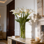 Cream calla lilies in a clear glass vase on a wood console beside a lit candle
