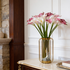 Pink and white calla lily stems in an amber glass vase by a stone fireplace