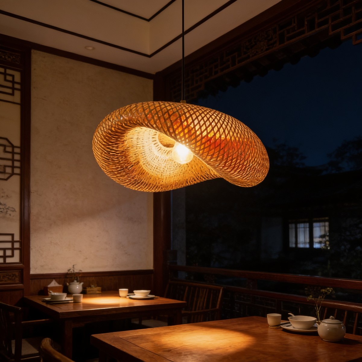 Saucer shaped woven pendant lit above wooden tables beside tall night windows