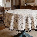 Round cream tablecloth with navy floral print and black tassel trim in a farmhouse room