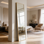 Slim gold framed floor mirror leaning by a living room window above a pale rug