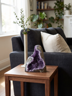 The amethyst cluster stands on a small walnut side table beside a dark sofa and a stem vase.