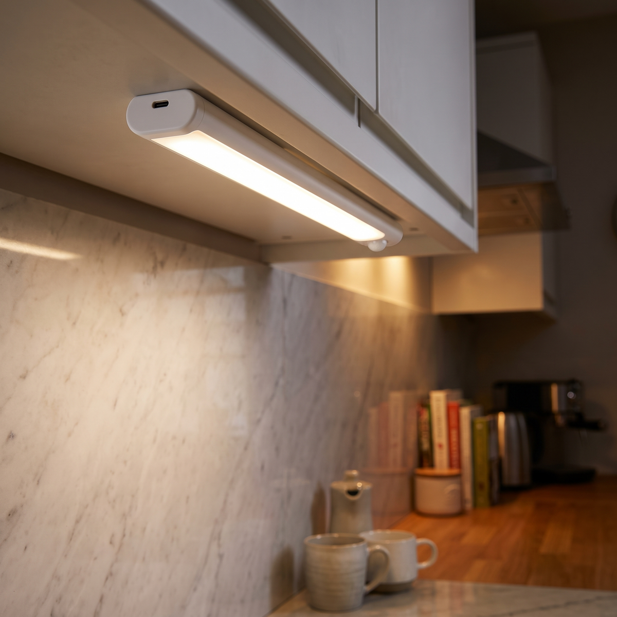 White sealed LED light bar with a round sensor lens under a cabinet, lighting a pale marble splashback and two mugs.