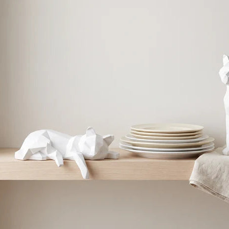 Faceted white cat lying on a shelf beside a stack of plain dinner plates