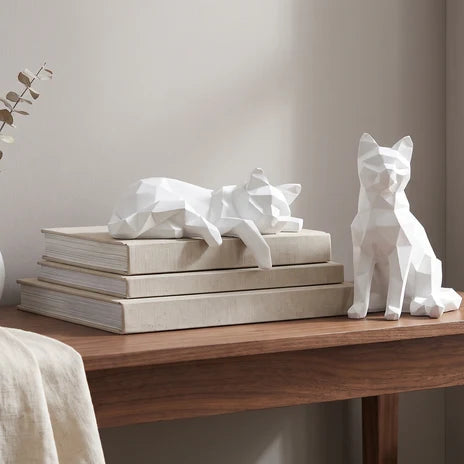 Two white geometric cat figures on a wood table, one resting on a stack of books